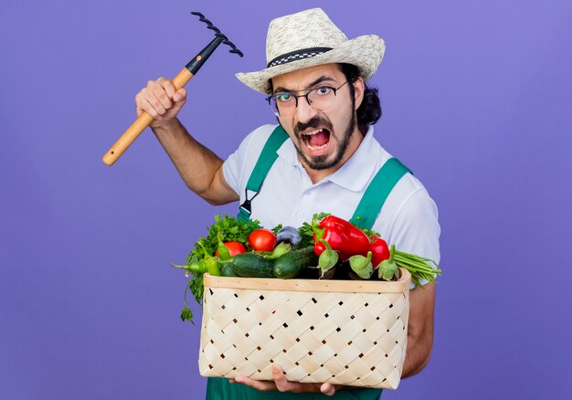 Young bearded gardener man wearing jumpsuit and hat holding crate full of vegetables swinging a mini rake with angry face standing over blue wall