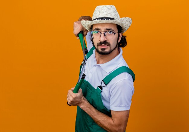 Young bearded gardener man wearing jumpsuit and hat holding hedge clippers looking at front worried standing over orange wall