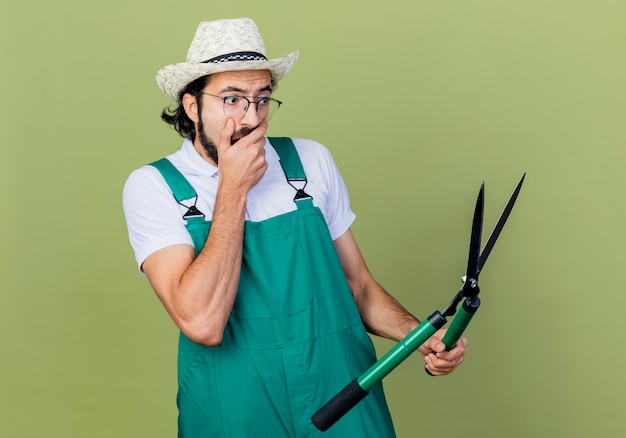 Молодой бородатый садовник в комбинезоне и шляпе с ножницами для живой изгороди выглядит удивленным и удивленным, стоя у светло-зеленой стены