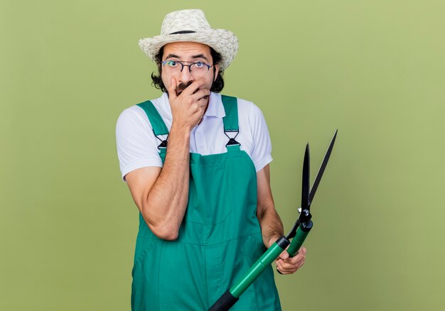 Young bearded gardener man wearing jumpsuit and hat holding hedge clippers looking surprised and amazed standing over light green wall