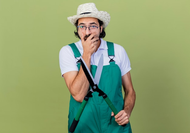 Young bearded gardener man wearing jumpsuit and hat holding hedge clippers looking surprised and amazed standing over light green wall