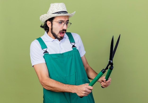Young bearded gardener man wearing jumpsuit and hat holding hedge clippers looking surprised and confused standing over light green wall