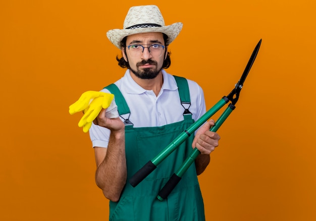 Young bearded gardener man wearing jumpsuit and hat holding hedge clippers and rubber gloves looking at front with sad expression standing over orange wall