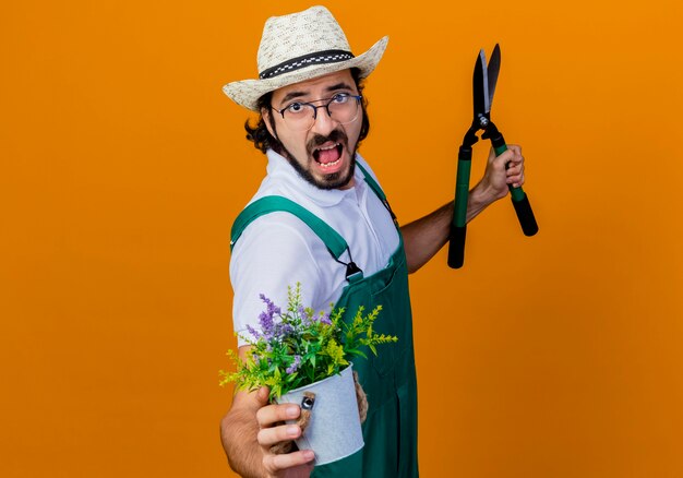 Молодой бородатый садовник в комбинезоне и шляпе держит ножницы для живой изгороди, показывая растение в горшке, кричащее разочарованное, стоящее над оранжевой стеной