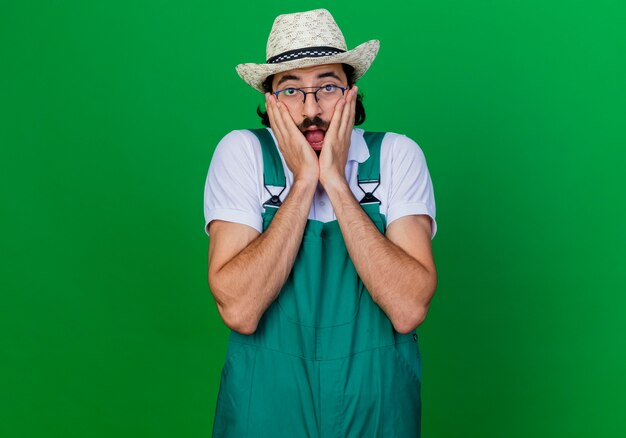 Young bearded gardener man wearing jumpsuit and hat holding his face with hands being shocked 