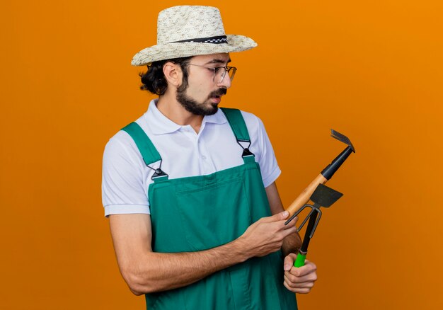 Young bearded gardener man wearing jumpsuit and hat holding mattock and mini rake looking at them being confused