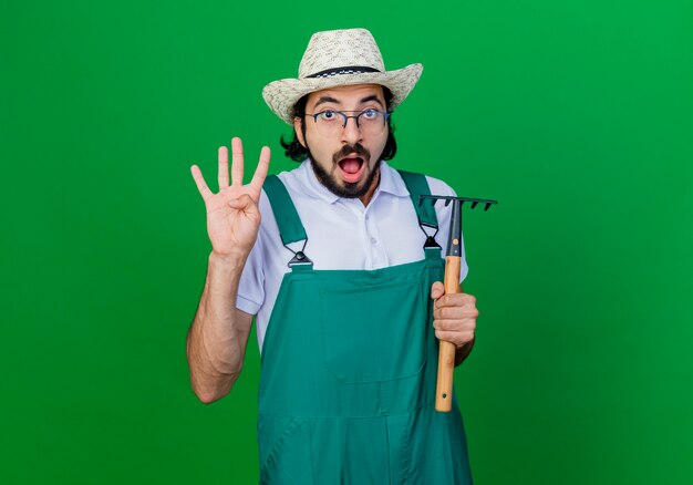 Young bearded gardener man wearing jumpsuit and hat holding mini rake being confused and surprised showing numberfour 