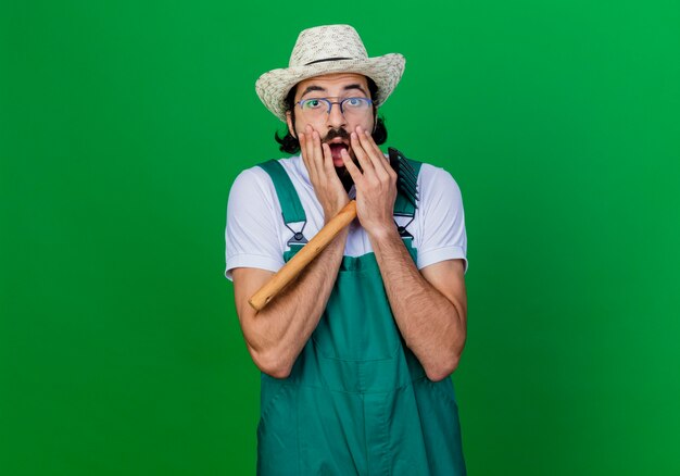 Молодой бородатый садовник в комбинезоне и шляпе с мини-граблями прикрывает рот руками, находясь в шоке