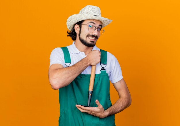 Young bearded gardener man wearing jumpsuit and hat holding mini rake looking at front smiling cheerfully standing over orange wall