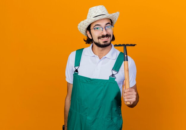 Young bearded gardener man wearing jumpsuit and hat holding mini rake looking at front smiling cheerfully standing over orange wall