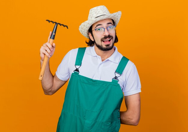 Young bearded gardener man wearing jumpsuit and hat holding mini rake looking at front smiling friendly standing over orange wall