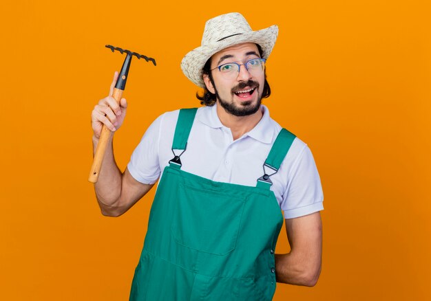 Молодой бородатый садовник в комбинезоне и шляпе, держащий мини-грабли, смотрит вперед, дружелюбно улыбаясь, стоя над оранжевой стеной