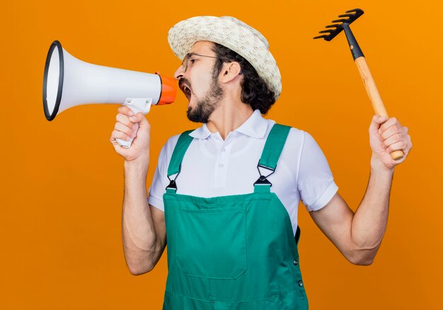 Young bearded gardener man wearing jumpsuit and hat holding mini rake shouting to megaphone being angry standing over orange wall