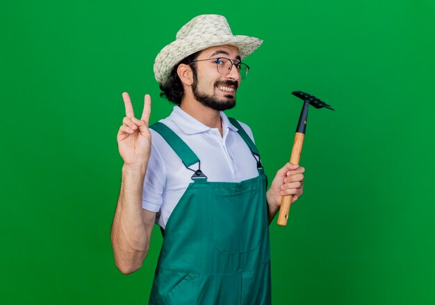 Young bearded gardener man wearing jumpsuit and hat holding mini rake smiling showing v- sign 