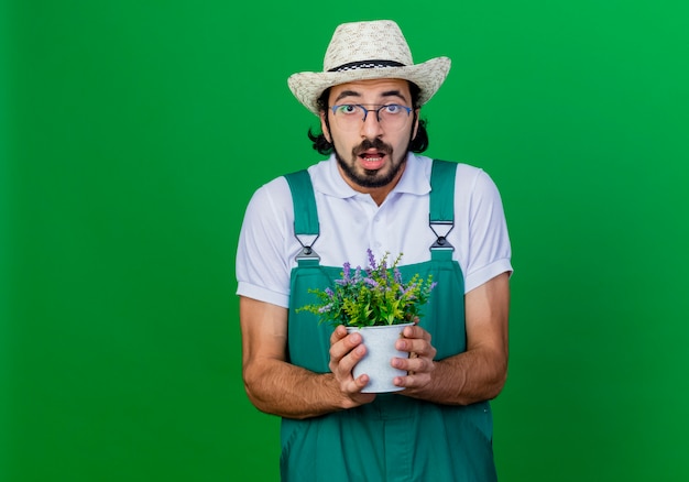 びっくりしている鉢植えの植物を保持しているジャンプスーツと帽子を身に着けている若いひげを生やした庭師の男