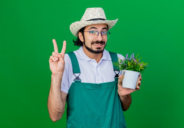 Молодой бородатый садовник в комбинезоне и шляпе держит горшечное растение с v-образным знаком