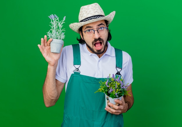 混乱して驚いている鉢植えの植物を保持しているジャンプスーツと帽子を身に着けている若いひげを生やした庭師の男