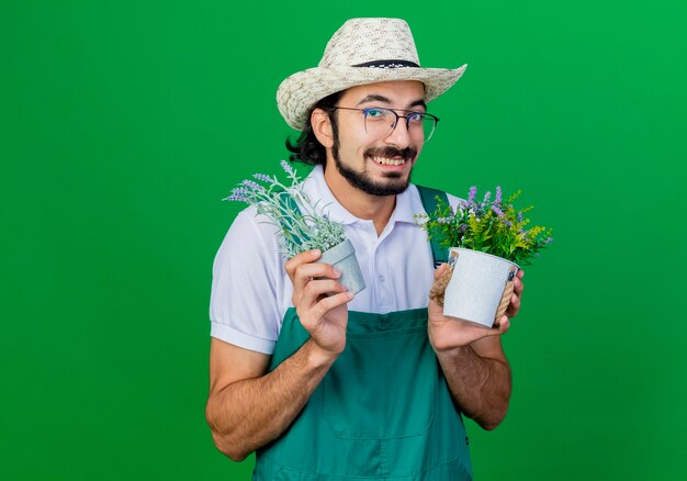 Young bearded gardener man wearing jumpsuit and hat holding potted plants smiling cheerfully 