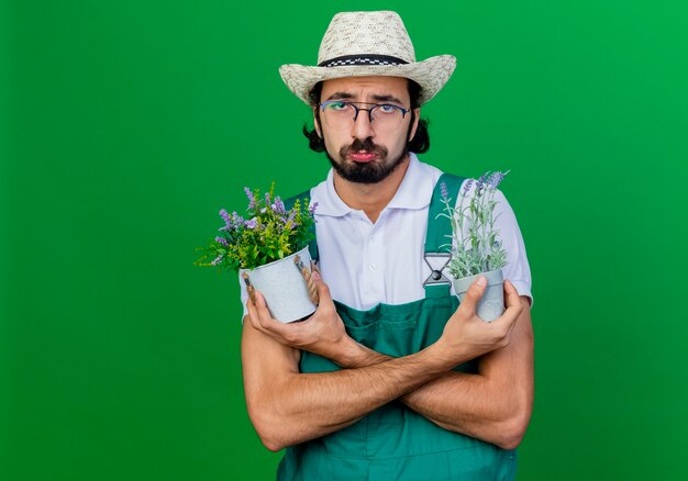 悲しい表情で鉢植えの植物を保持しているジャンプスーツと帽子を身に着けている若いひげを生やした庭師の男