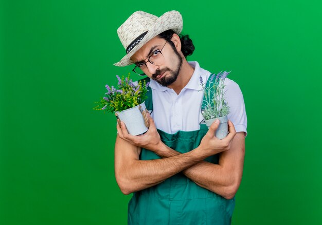 つなぎ服と帽子をかぶって鉢植えの植物を持っている若いひげを生やした庭師の男