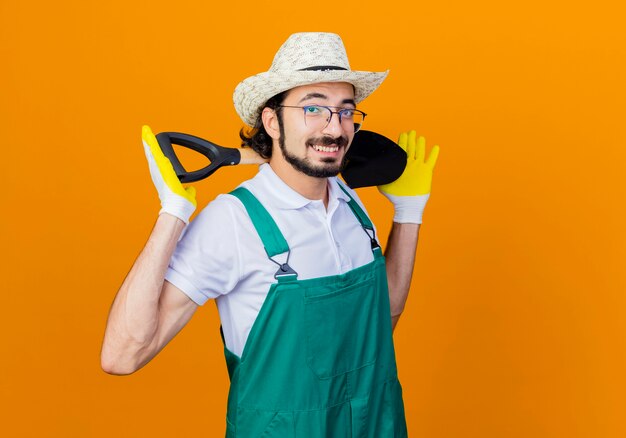 젊은 수염 정원사 남자 점프 슈트와 모자를 쓰고 삽을 들고 오렌지 벽 위에 유쾌하게 서 웃고 앞을보고