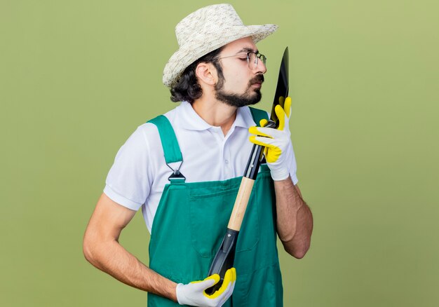 Young bearded gardener man wearing jumpsuit and hat holding shovel looking at it closely standing over light green wall