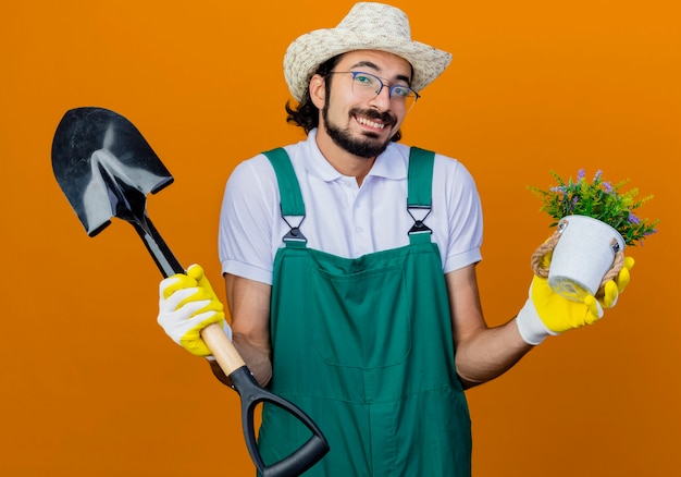 Young bearded gardener man wearing jumpsuit and hat holding shovel and potted plant looking confused shrugging shoulders 