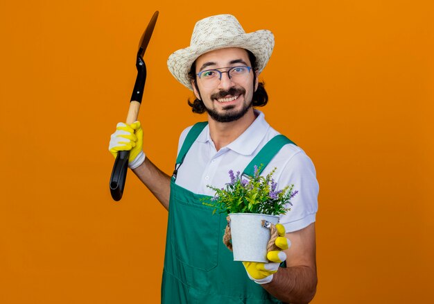 Молодой бородатый садовник в комбинезоне и шляпе держит лопату и горшечное растение, глядя вперед, улыбаясь, стоя над оранжевой стеной