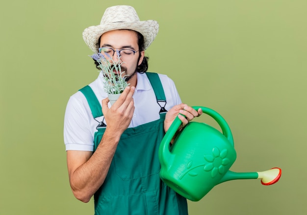 Giovane giardiniere barbuto uomo che indossa tuta e cappello azienda annaffiatoio e pianta in vaso inalazione di odore gradevole in piedi sopra il muro verde chiaro