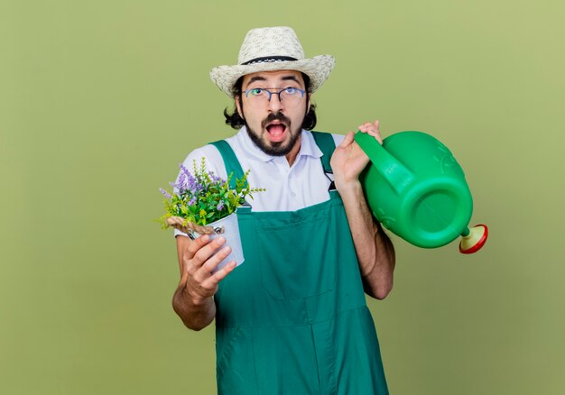 じょうろと鉢植えの植物を持ってジャンプスーツと帽子をかぶった若いひげを生やした庭師の男は、薄緑色の壁の上に立って驚いて驚いています