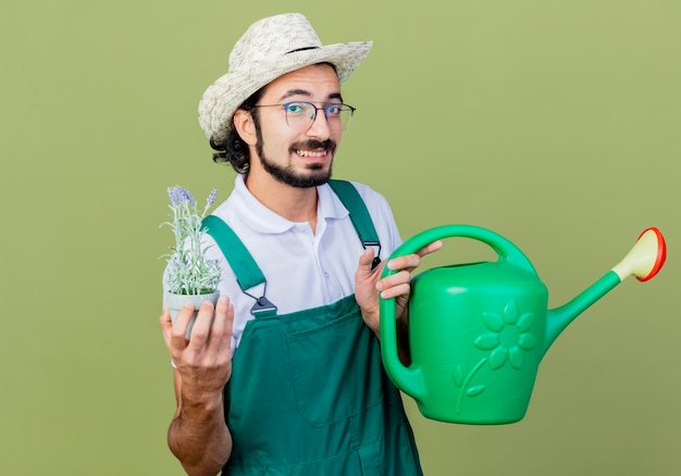 Giovane giardiniere barbuto uomo che indossa tuta e cappello tenendo annaffiatoio e pianta in vaso guardando la parte anteriore sorridente con la faccia felice in piedi sopra la parete verde chiaro