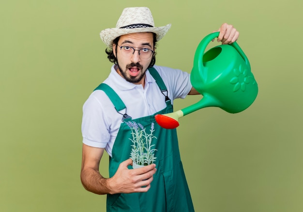 Giovane giardiniere barbuto uomo che indossa tuta e cappello tenendo annaffiatoio e pianta in vaso guardando davanti sorpreso in piedi sopra il muro verde chiaro