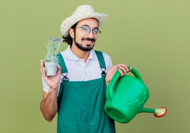 Young bearded gardener man wearing jumpsuit and hat holding watering can and potted plant smiling with happy face 