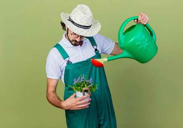 Young bearded gardener man wearing jumpsuit and hat holding watering can and potted plant watering it standing over light green wall
