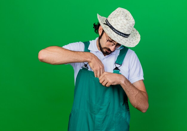 Young bearded gardener man wearing jumpsuit and hat looking down putting something into his pocket 