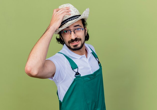 薄緑色の壁の上に立っている彼の帽子に触れて笑顔で正面を見てジャンプスーツと帽子を身に着けている若いひげを生やした庭師の男