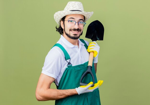 Молодой бородатый садовник в комбинезоне и шляпе, представляя лопату, глядя вперед, улыбаясь, стоя над светло-зеленой стеной