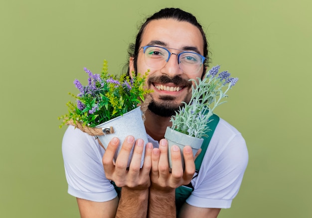 Молодой бородатый садовник в комбинезоне держит горшечные растения, глядя вперед, весело улыбаясь, стоя над светло-зеленой стеной
