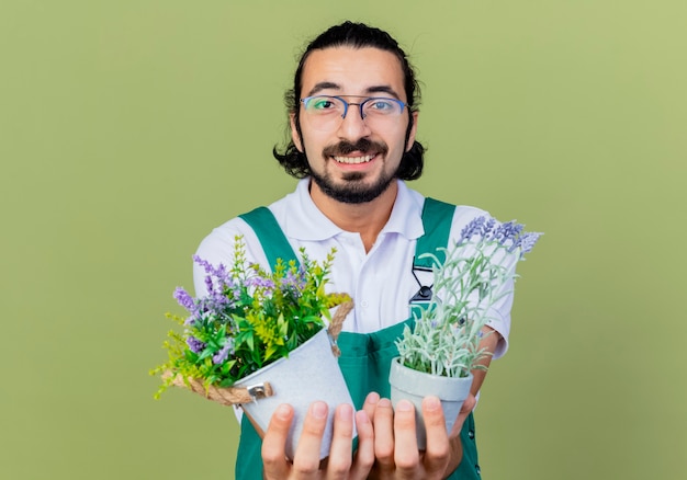 Free photo young bearded gardener man wearing jumpsuit holding potted plants looking at front smiling cheerfully standing over light green wall