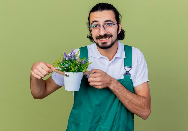 Young bearded gardener man wearing jumpsuit showing potted plant smiling looking at front standing over light green wall