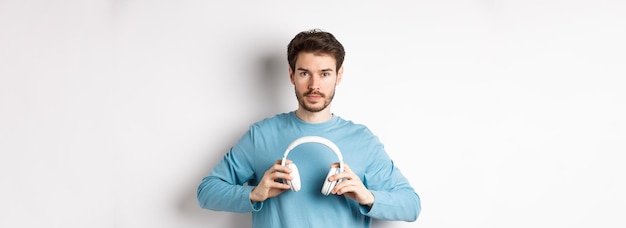 Young bearded guy in blue sweatshirt put on wireless headphones listening music standing on white ba