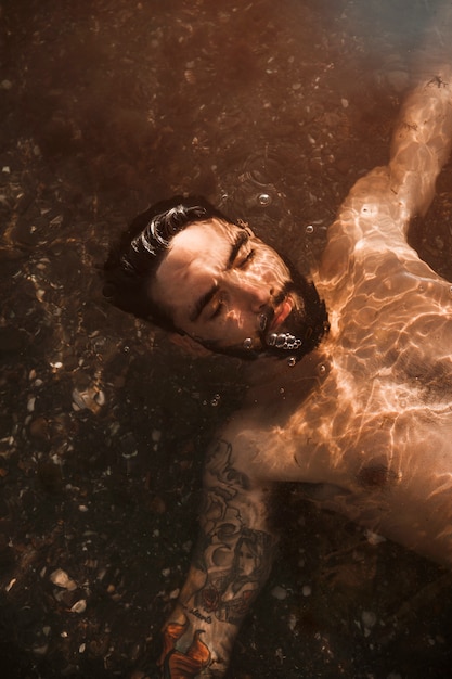 Young bearded guy lying under water on shore