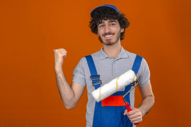 Young bearded handsome builder wearing construction uniform and cap holding paint roller pointing at something behind him smiling over isolated orange wall