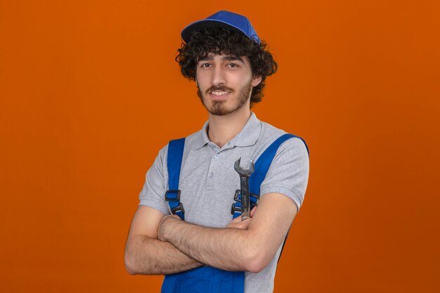 Free photo young bearded handsome builder wearing construction uniform and cap standing with arms crossed self-confident smiling over isolated orange wall