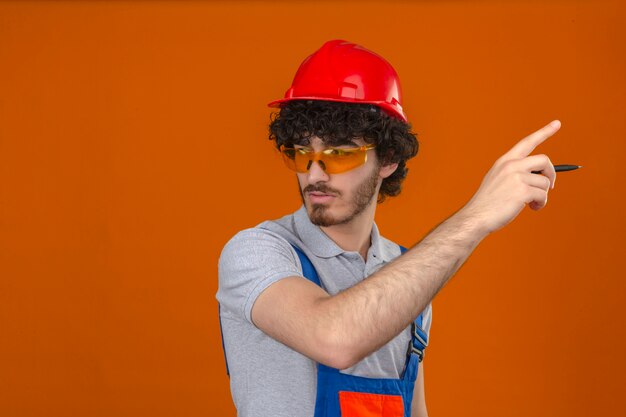 Young bearded handsome builder wearing construction uniform glasses and safety helmet looking aside holding pen in hand and pointing with serious face over isolated orange wall