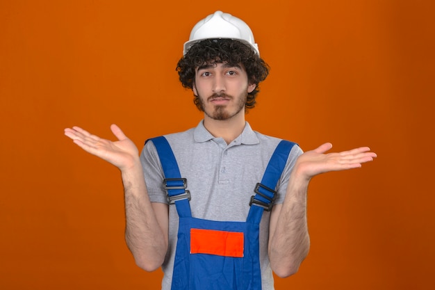 Young bearded handsome builder wearing construction uniform and safety helmet clueless and confused expression with arms and hands raised having doubts over isolated orange wall