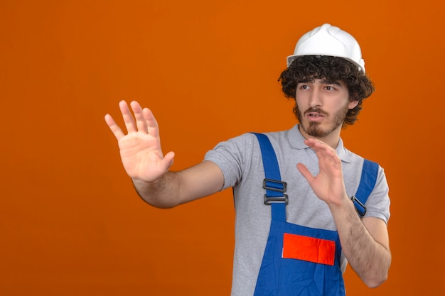 Young bearded handsome builder wearing construction uniform and safety helmet holding his hands up telling do not come closer over isolated orange wall