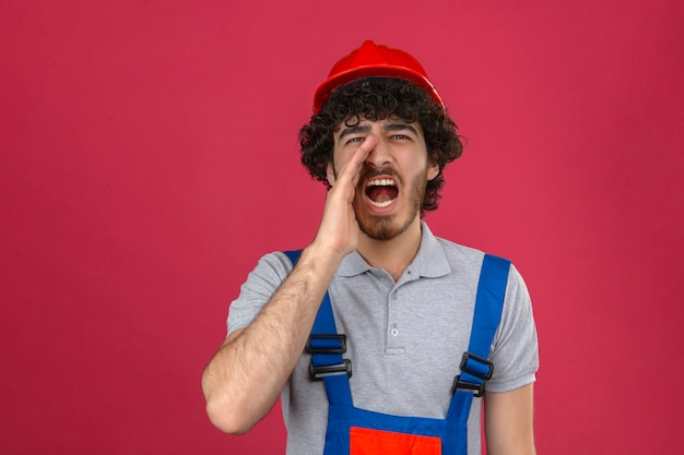 Young bearded handsome builder wearing construction uniform and safety helmet shouting and holding palm near opened mouth over isolated pink wall