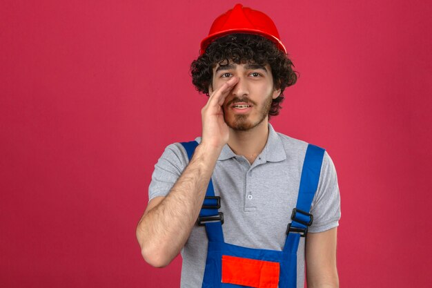 Young bearded handsome builder wearing construction uniform and safety helmet shouting with palm near mouth over isolated pink wall