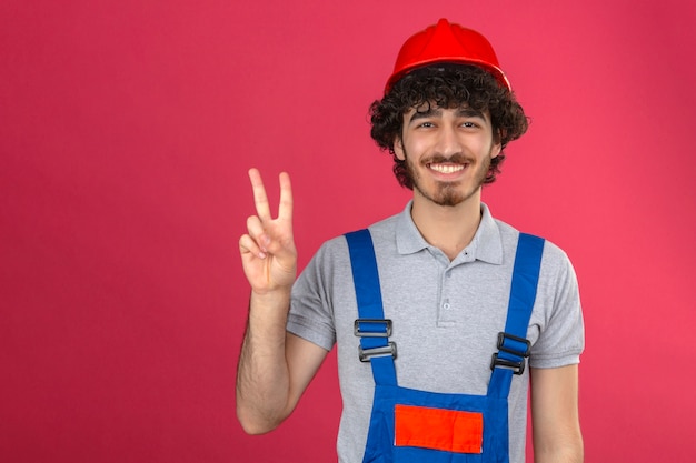 Young bearded handsome builder wearing construction uniform and safety helmet smiling cheerfully showing victory sign over isolated pink wall